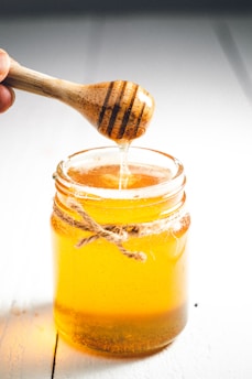 A wooden honey dipper is lifting honey from a glass jar filled with golden honey. The jar is tied with a rustic string around its neck and sits on a white surface.