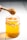 A wooden honey dipper is lifting honey from a glass jar filled with golden honey. The jar is tied with a rustic string around its neck and sits on a white surface.