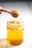 A wooden honey dipper is lifting honey from a glass jar filled with golden honey. The jar is tied with a rustic string around its neck and sits on a white surface.