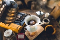 A cozy coffee-making setup featuring a pour-over method. A metallic kettle is pouring hot water onto ground coffee in a white dripper, which sits atop a glass container. Surrounding the setup are various items including a gas stove, a coffee mug, a jar, and what appears to be a sliced fruit.