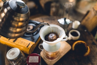 A cozy coffee-making setup featuring a pour-over method. A metallic kettle is pouring hot water onto ground coffee in a white dripper, which sits atop a glass container. Surrounding the setup are various items including a gas stove, a coffee mug, a jar, and what appears to be a sliced fruit.