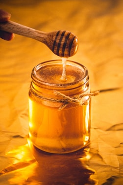 A glass jar filled with golden honey, sitting on a reflective golden surface. A wooden honey dipper is dripping honey into the jar. The jar is wrapped with a piece of thick twine.