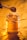 A glass jar filled with golden honey, sitting on a reflective golden surface. A wooden honey dipper is dripping honey into the jar. The jar is wrapped with a piece of thick twine.