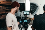 Two men are engaged in a video production setting. One man operates a professional camera and monitors the footage on a screen, while the other appears to be holding a smartphone, possibly assisting or recording as well. The background features photography equipment, including a large softbox light and a tripod.