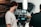 Two men are engaged in a video production setting. One man operates a professional camera and monitors the footage on a screen, while the other appears to be holding a smartphone, possibly assisting or recording as well. The background features photography equipment, including a large softbox light and a tripod.