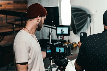 Two men are engaged in a video production setting. One man operates a professional camera and monitors the footage on a screen, while the other appears to be holding a smartphone, possibly assisting or recording as well. The background features photography equipment, including a large softbox light and a tripod.