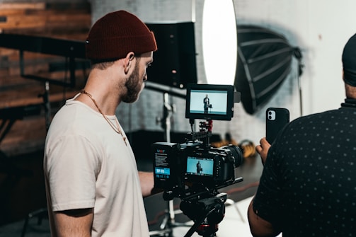 Two men are engaged in a video production setting. One man operates a professional camera and monitors the footage on a screen, while the other appears to be holding a smartphone, possibly assisting or recording as well. The background features photography equipment, including a large softbox light and a tripod.