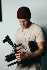 A person wearing a brown beanie and a light beige shirt is holding a professional camera rig. The camera appears to be equipped with additional gear, suggesting a focus on filmmaking or video production.