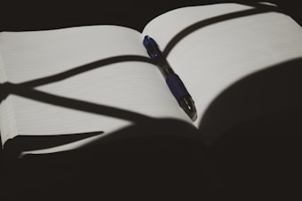An open notebook with lined pages is placed under dramatic lighting, casting shadows across the pages. A pen with a blue grip rests diagonally on the notebook.