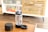 A transparent cylindrical device with a black top and a small bowl, likely a water pipe or bong, is placed on a wooden table. Next to it is a round, black container with a lid and a coaster. The background shows a wooden cabinet with slats and colorful books on a white shelf.