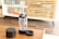 A transparent cylindrical device with a black top and a small bowl, likely a water pipe or bong, is placed on a wooden table. Next to it is a round, black container with a lid and a coaster. The background shows a wooden cabinet with slats and colorful books on a white shelf.