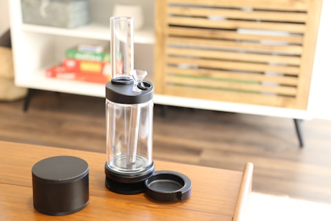 A transparent cylindrical device with a black top and a small bowl, likely a water pipe or bong, is placed on a wooden table. Next to it is a round, black container with a lid and a coaster. The background shows a wooden cabinet with slats and colorful books on a white shelf.