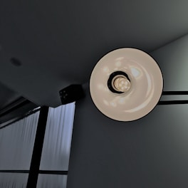 A minimalist architectural setting featuring a ceiling light fixture with a circular design, positioned against a gray background. The light bulb is centered within a white, reflective disc, casting a soft glow. Adjacent to the fixture are large windows with vertically aligned blinds, allowing diffused natural light into the space.