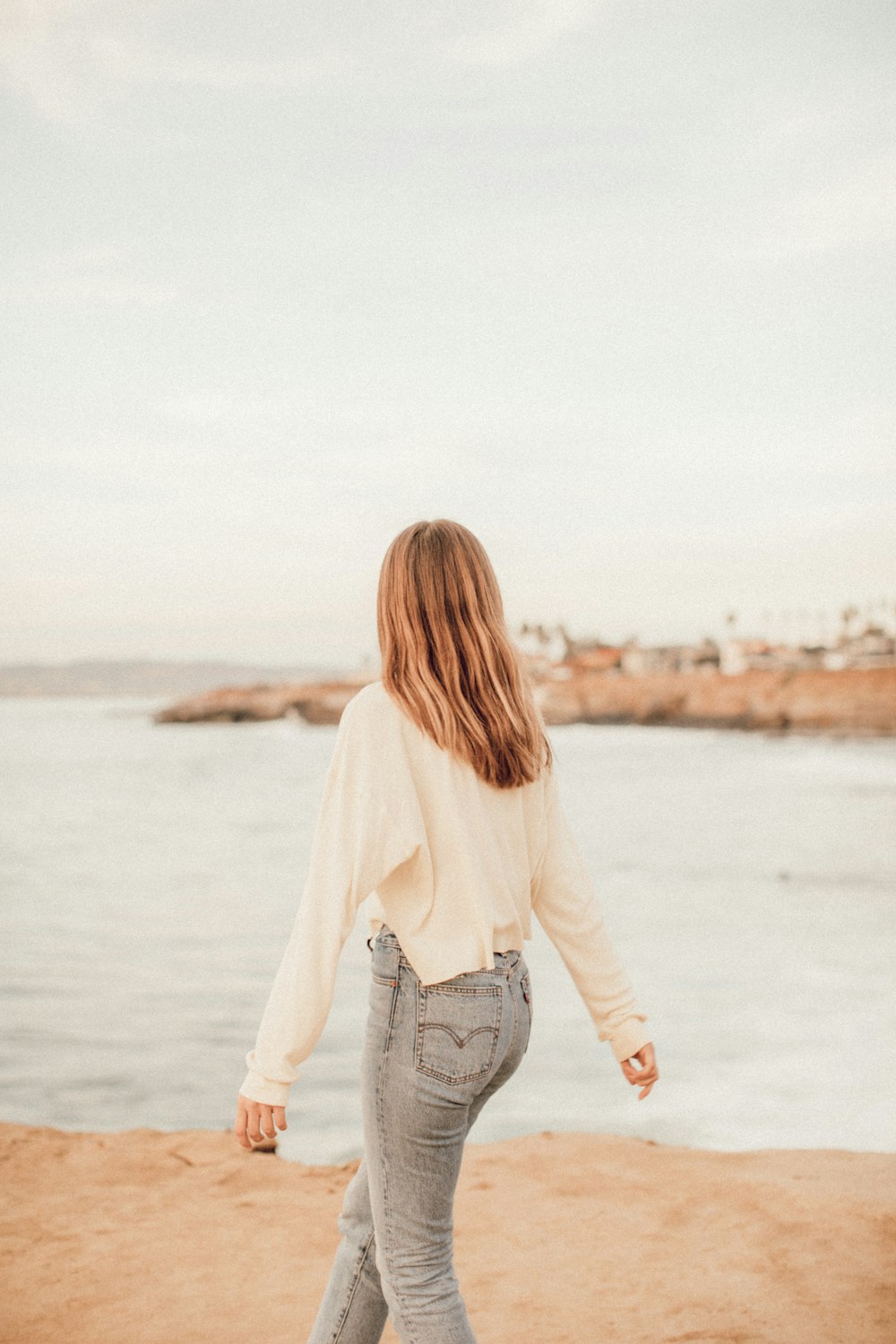 흰색 긴팔 셔츠와 파란색 데님 청바지를 입은 여자가 낮 동안 해변에 서 있다