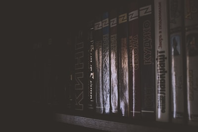 A dimly lit bookshelf with a row of books lined up, showing mainly the spines of the books which are mostly dark with some reflective surfaces. The lighting creates a moody and mysterious atmosphere.