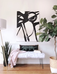 A modern interior space featuring a large abstract black and white painting on the wall. Below the painting, a bench with a fluffy white cushion is adorned with a patterned pillow featuring a stylized animal design, and a pale pink blanket draped over it. Two books are stacked next to the pillow. A tall floor lamp with a white shade stands on the left, while potted plants, including a snake plant and a large leafy plant in a woven basket, add greenery to the setting. The flooring is wooden, and the walls are painted white.