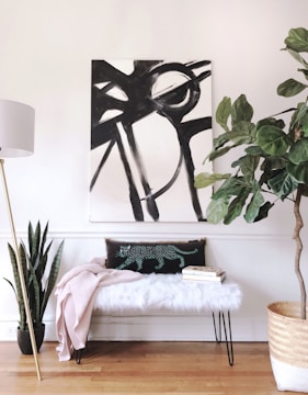 A modern interior space featuring a large abstract black and white painting on the wall. Below the painting, a bench with a fluffy white cushion is adorned with a patterned pillow featuring a stylized animal design, and a pale pink blanket draped over it. Two books are stacked next to the pillow. A tall floor lamp with a white shade stands on the left, while potted plants, including a snake plant and a large leafy plant in a woven basket, add greenery to the setting. The flooring is wooden, and the walls are painted white.