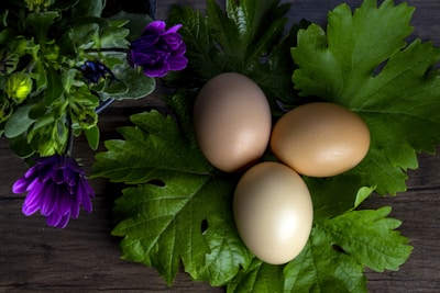 Three brown eggs are placed on a bed of large, green leaves on a wooden surface. Surrounding the eggs is a cluster of purple flowers with lush green foliage.
