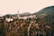 A small village is nestled in a mountainous landscape. The village features a prominent mosque with a tall minaret, surrounded by houses scattered across the hillside. The vegetation appears lush, and the hills in the background create a picturesque setting.