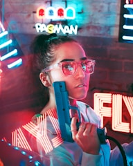 A person in stylish neon glasses is holding a retro arcade gun. Colorful neon signs in the background, including a Pac-Man sign, create a vibrant, futuristic atmosphere.