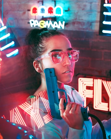 A person in stylish neon glasses is holding a retro arcade gun. Colorful neon signs in the background, including a Pac-Man sign, create a vibrant, futuristic atmosphere.