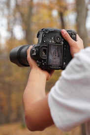 A person is holding a DSLR camera with a long lens, capturing an image of an autumn forest, visible on the camera's display screen. The background displays a blur of fall foliage, predominantly in warm tones.
