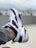 A white and black sneaker with red accents is prominently presented. The shoe features a distinctive red swoosh logo, and appears to be worn on a person's foot wearing gray pants. The background is blurred, suggesting an outdoor setting.