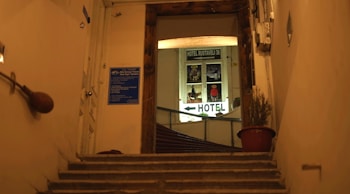 A dimly lit staircase leading to a hotel entrance with a visible sign that reads 'Hotel Rustaveli 36'. The walls are beige, and there is a blue information plaque on the left side. A potted plant in a red pot is situated on the right side of the stairs.