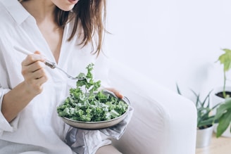woman in white dress holding silver fork and knife slicing green vegetable on white ceramic bowl