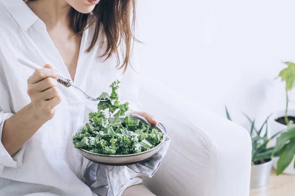 Mujer en vestido blanco sosteniendo tenedor de plata y cuchillo cortando vegetales verdes en cuenco de cerámica blanca