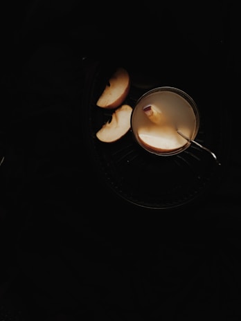A cup containing a light-colored beverage with a slice of apple floating on top and a spoon inserted into the cup. Sliced apples are placed beside the cup, all set against a dark background.