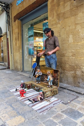 A street performer operates a puppet show on a city sidewalk. The puppets are dressed in colorful clothing and appear to be musical band members, with instruments like a guitar and a drum. The performer stands beside a backdrop of a stone wall and a storefront with signs in the window.
