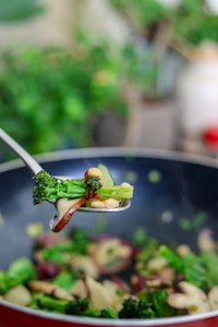 A close-up of a spoon holding sautéed vegetables, including broccoli, red peppers, and mushrooms, over a pan filled with more vegetables. The background is blurred, highlighting the freshness and colors of the ingredients.