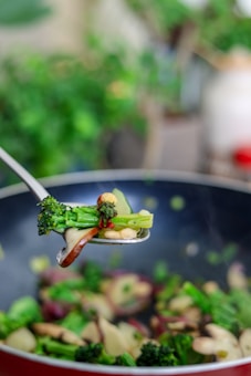 A close-up of a spoon holding saut&eacute;ed vegetables, including broccoli, red peppers, and mushrooms, over a pan filled with more vegetables. The background is blurred, highlighting the freshness and colors of the ingredients.