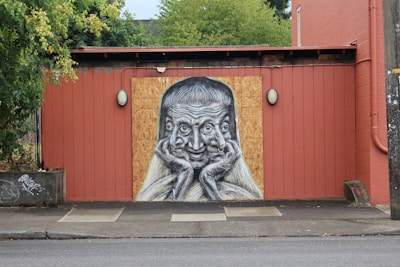 A mural on a red wooden wall depicts a monochrome surrealistic face with multiple eyes and hands clasped together under the chin. The surrounding area includes greenery and a cement sidewalk with some graffiti on the left corner.