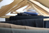 A close-up view of multiple pairs of trousers hanging on wooden hangers, arranged in a row. The hangers are spaced evenly, displaying a range of dark and neutral colors such as black, navy, and brown.