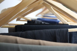 A close-up view of multiple pairs of trousers hanging on wooden hangers, arranged in a row. The hangers are spaced evenly, displaying a range of dark and neutral colors such as black, navy, and brown.