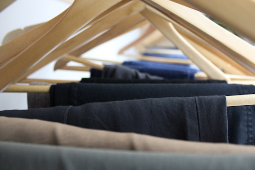 A close-up view of multiple pairs of trousers hanging on wooden hangers, arranged in a row. The hangers are spaced evenly, displaying a range of dark and neutral colors such as black, navy, and brown.