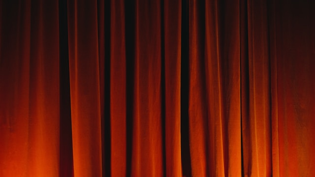 Dark orange and brown theater curtains with subtle lighting that creates shadows and highlights on the fabric. The heavy drapery appears plush and conveys a sense of anticipation.