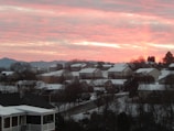 A residential neighborhood with houses covered in snow, set against a sunset with vibrant pink and orange colors. The landscape includes distant mountains and numerous bare trees scattered among the homes.