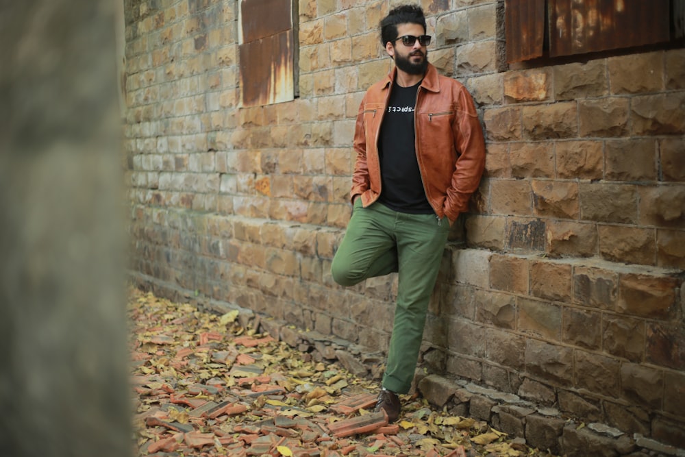 man in brown leather jacket and green pants leaning on brown brick wall during daytime