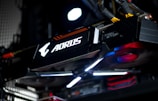 A close-up view of a high-performance graphics card inside a computer, featuring prominent branding and illuminated components. The cables and surrounding hardware components are partially visible, contributing to a sleek, modern tech aesthetic.