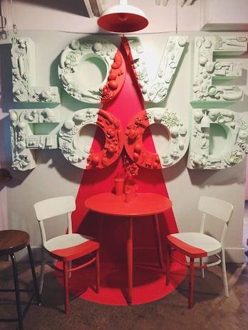 A decorative installation features the words 'LOVE FOOD' in large, bold letters, embellished with various food-related items. The letters are divided into sections of light blue and red, creating a vivid contrast. A small round table, painted red, is flanked by two chairs with alternating white and red sections. A pendant light colored in red hangs above, focusing on the table.