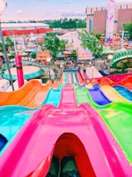 A vibrant and colorful view from the top of a giant slide at a fairground. The slide is made up of lanes in bright pink, blue, green, and orange. In the background, an array of carnival rides, food stands, and games are visible with people walking around and enjoying the fair. Buildings and trees surround the fair in the distance under a clear sky.
