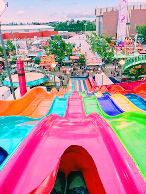 A vibrant and colorful view from the top of a giant slide at a fairground. The slide is made up of lanes in bright pink, blue, green, and orange. In the background, an array of carnival rides, food stands, and games are visible with people walking around and enjoying the fair. Buildings and trees surround the fair in the distance under a clear sky.