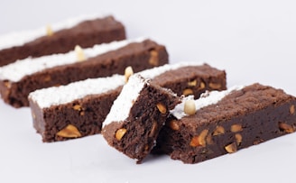Several chocolate brownies are arranged in a line, featuring a dusting of powdered sugar on top and pieces of nuts embedded inside. The brownies are stacked in a way that one piece rests on another, showcasing their rich texture.