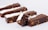 Several chocolate brownies are arranged in a line, featuring a dusting of powdered sugar on top and pieces of nuts embedded inside. The brownies are stacked in a way that one piece rests on another, showcasing their rich texture.