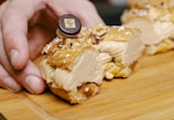 A close-up view of a pastry being held by a hand on a wooden surface. The pastry is filled with a creamy filling and topped with a decorative chocolate piece. The texture of the pastry shell is flaky and golden brown.