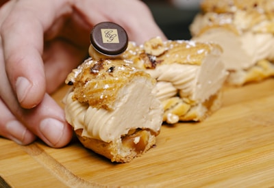 A close-up view of a pastry being held by a hand on a wooden surface. The pastry is filled with a creamy filling and topped with a decorative chocolate piece. The texture of the pastry shell is flaky and golden brown.