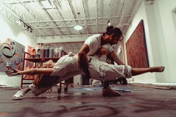 Two dancers are performing an intricate lift in an art studio with a high ceiling and various paintings and art supplies around. The woman is arched backward, balancing on the man's knee, while he supports her with his arms. The environment suggests a creative and dynamic energy.